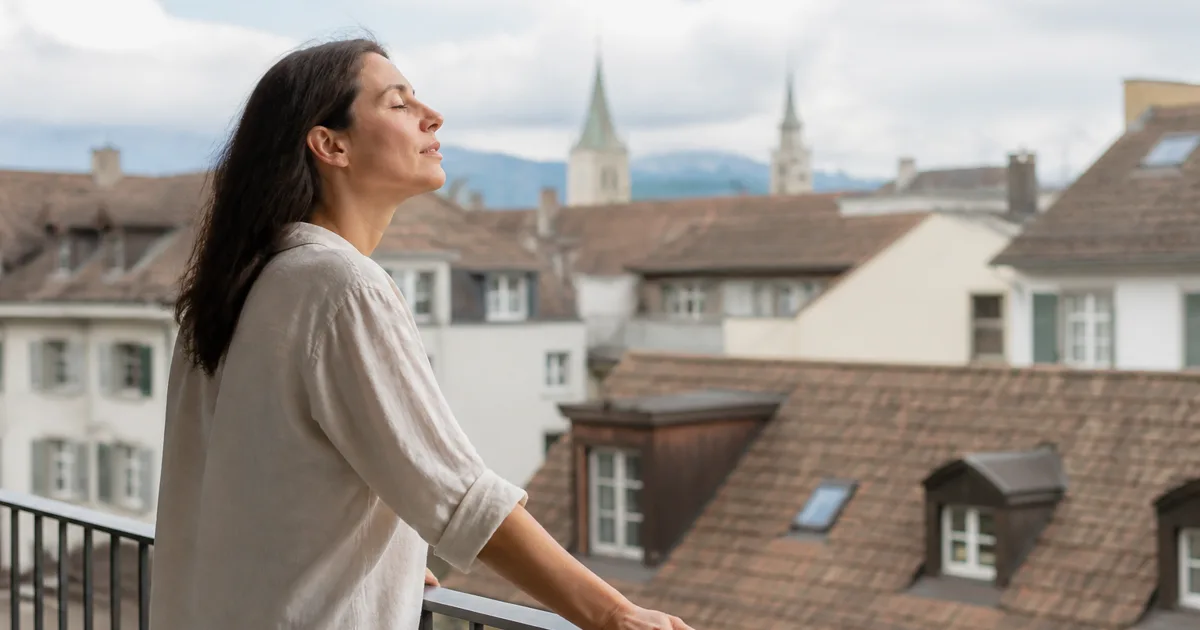 Person hält auf einem Balkon inne, beide Hände am Geländer, Schweizer Dächer dahinter